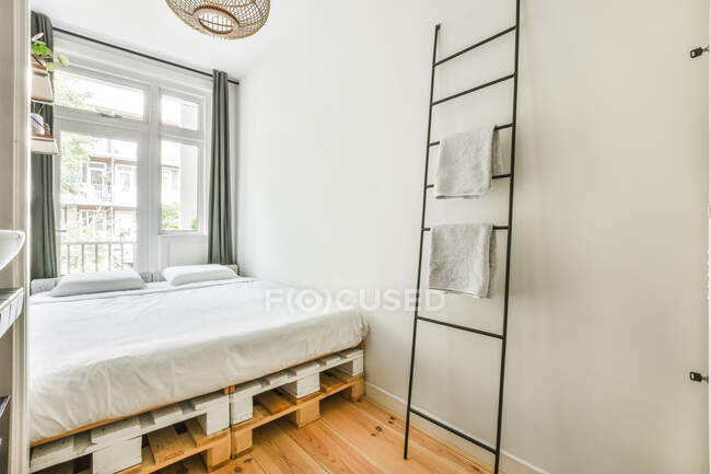 Escalera con toallas colocadas cerca de la cama de la plataforma en el dormitorio de estilo minimalista ligero con paredes grises en el día - foto de stock