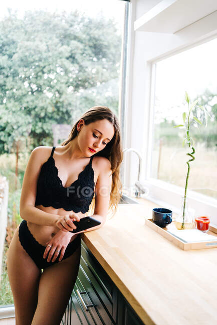 Seductive female in black lingerie and with red lips leaning at counter in kitchen at home and looking her mobile — Stock Photo