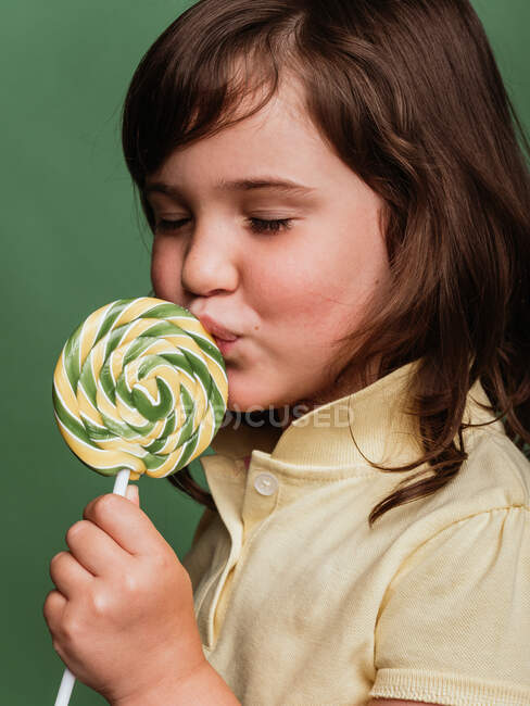 Funny preteen child licking sweet swirl lollipop on green background in studio with closed eyes — Stock Photo