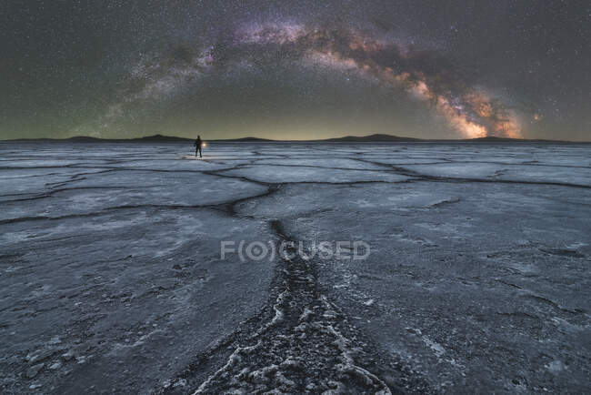 Distant silhouette of explorer standing and holding a flashlight in dry salt lagoon on background of starry sky with glowing Milky Way at night — Stock Photo
