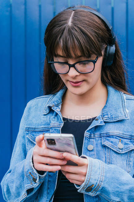 Positive female in headphones browsing on smartphone while standing on blue background in street — Stock Photo