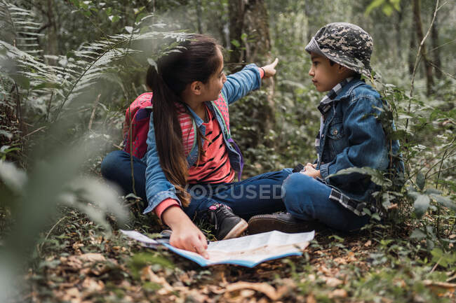 Chica étnica mostrando planta verde a hermano mientras mira hacia otro lado y sentado en el terreno con mapa de papel y lupa en los bosques - foto de stock
