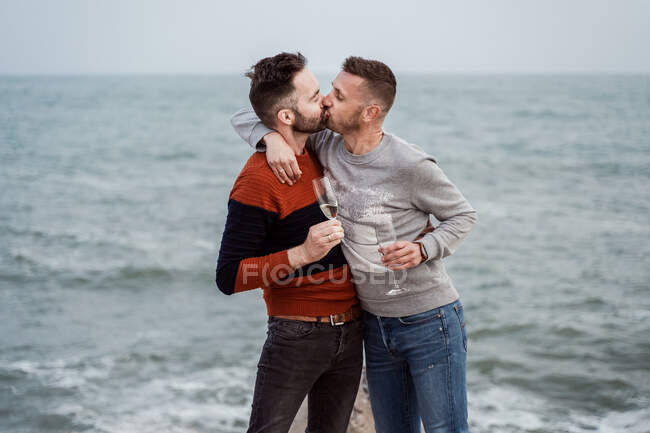 Homosexual male partners with modern haircuts enjoying champagne from glasses while kissing on ocean coast in daytime — Stock Photo