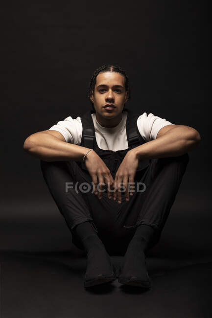 Confident young ethnic hipster male model with braided hair wearing white shirt with black clothes looking at camera while sitting in dark studio — Stock Photo
