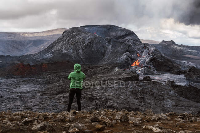 Обратный вид на неузнаваемого путешественника, стоящего на горе и созерцающего огненный Федрадальсфьолл в Исландии — стоковое фото