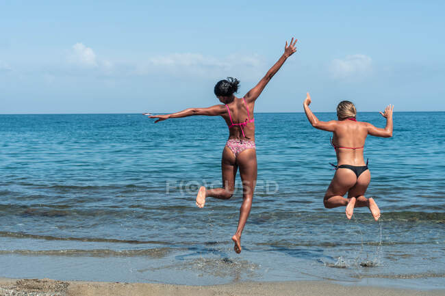 Back view of anonymous joyful diverse female tourists in swimwear jumping over ocean beach under cloudy blue sky — Stock Photo