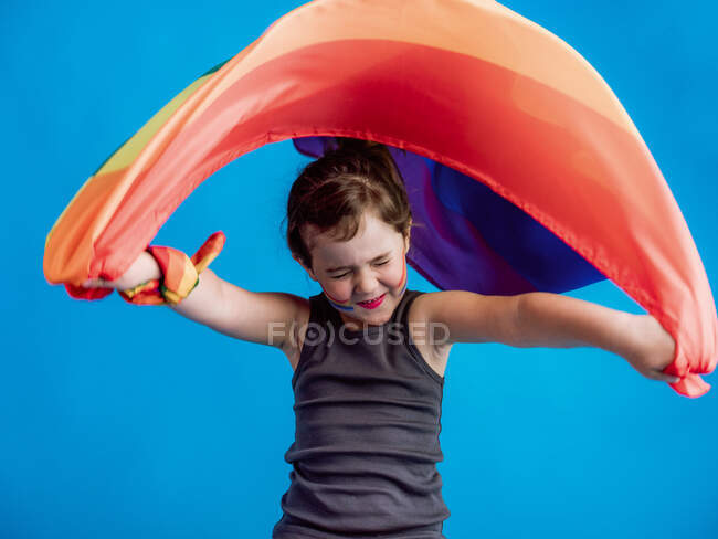 Adorável menina fechar os olhos ao levantar bandeira colorida acima da cabeça contra o fundo azul vibrante — Fotografia de Stock