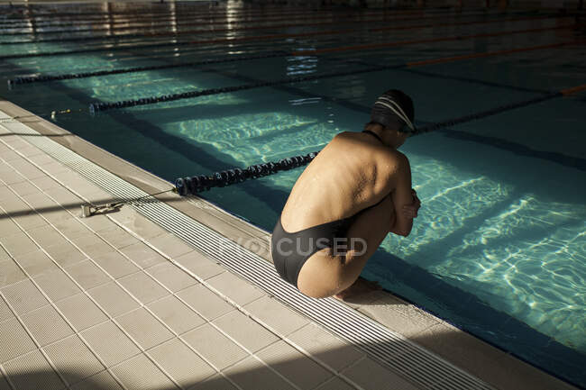 Young beautiful woman inside the indoor pool, wearing black swimsuit, squatting on the edge, lateral view — Stock Photo