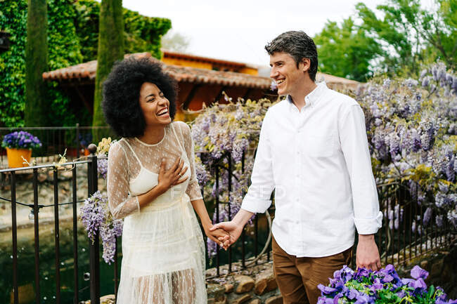 Zufriedenes multiethnisches Paar hält Händchen, während es im Garten mit blühenden lila Glyzinien spaziert und das gemeinsame Wochenende genießt — Stockfoto