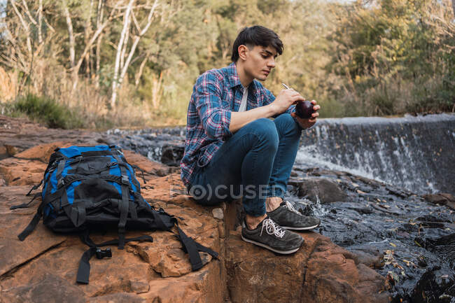 Senderista hombre sediento con mochila bebiendo agua de la taza con paja mientras está sentado en la roca cerca de la cascada en el bosque y mirar hacia adentro durante el descanso - foto de stock