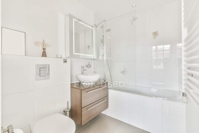 Interior elegante do banheiro branco com lavatório e armário de madeira perto da cabine do chuveiro com divisória de vidro no apartamento moderno — Fotografia de Stock