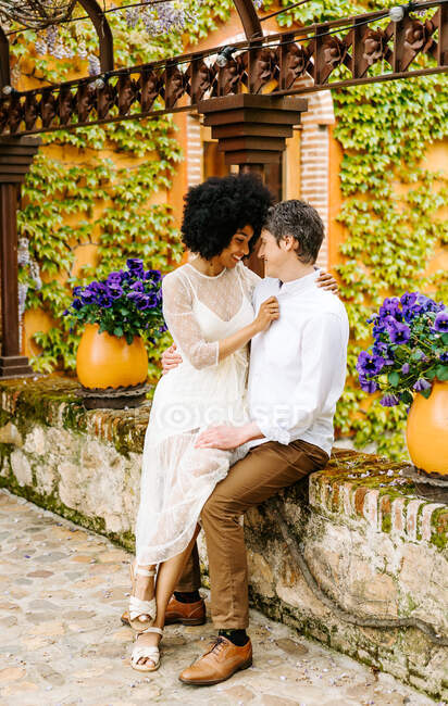 Cheerful African American woman sitting on knees of loving man while hugging on stone border in aged town — Stock Photo