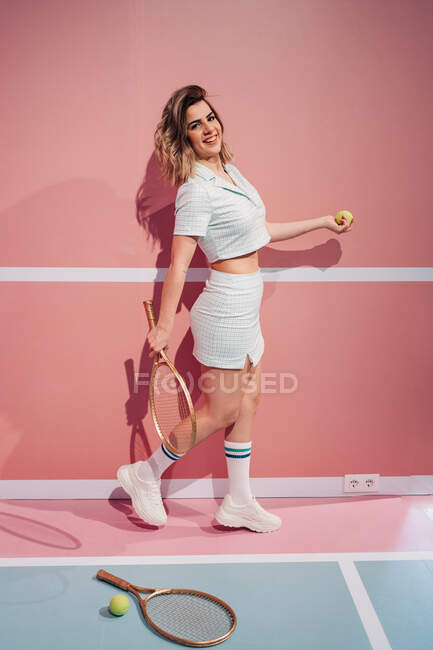 Вид сбоку на веселую молодую спортсменку в кроссовках с теннисной ракеткой, смотрящую в камеру на спортивной площадке — стоковое фото