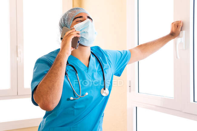 Médico masculino en uniforme y con estetoscopio de pie en el hospital y hablando en el teléfono inteligente mientras mira hacia arriba - foto de stock