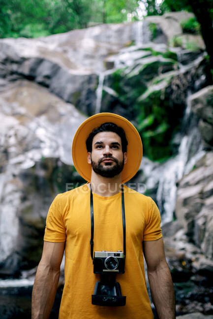 Male explorer in yellow clothes and with vintage photo camera standing on background of waterfall in woods and looking up — Stock Photo