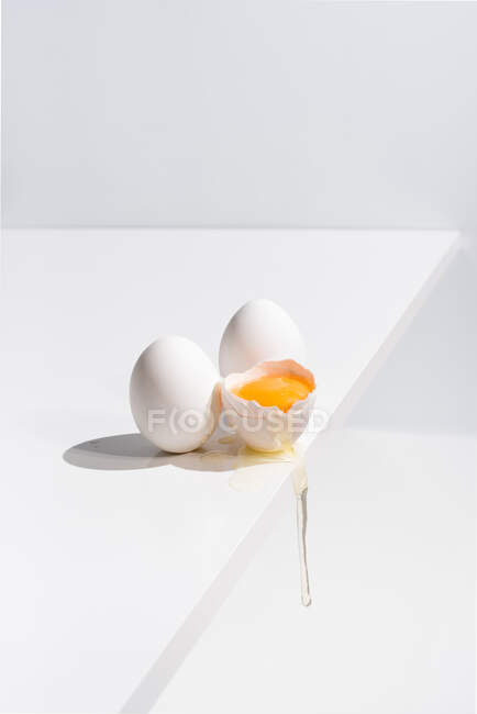 Alto angolo di uovo intero e rotto con tuorlo in guscio posto sul bordo del tavolo su sfondo bianco in studio — Foto stock