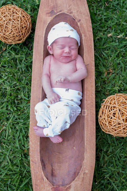 Vista superior de lindo bebé recién nacido durmiendo mientras está acostado en una bañera de madera colocada sobre hierba verde - foto de stock