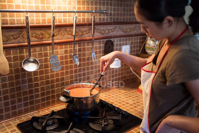 Сверху урожая женщина добавляет соль в кастрюлю во время приготовления соуса маринара из помидоров на плите на кухне — стоковое фото