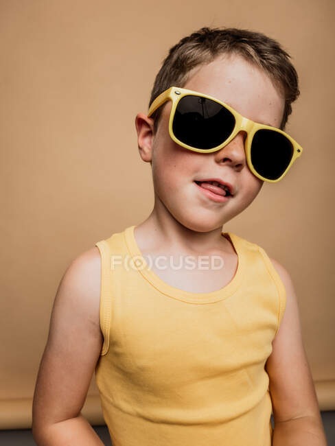 Cool menino pré-adolescente na moda óculos de sol amarelos mostrando língua e olhando para a câmera no fundo marrom em estúdio — Fotografia de Stock