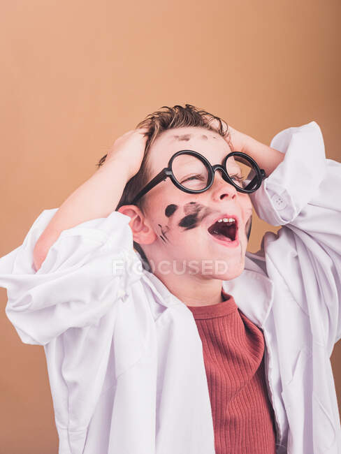 Chimico ragazzo in accappatoio da laboratorio e bicchieri di plastica guardando lontano su sfondo beige con le mani sulla testa — Foto stock