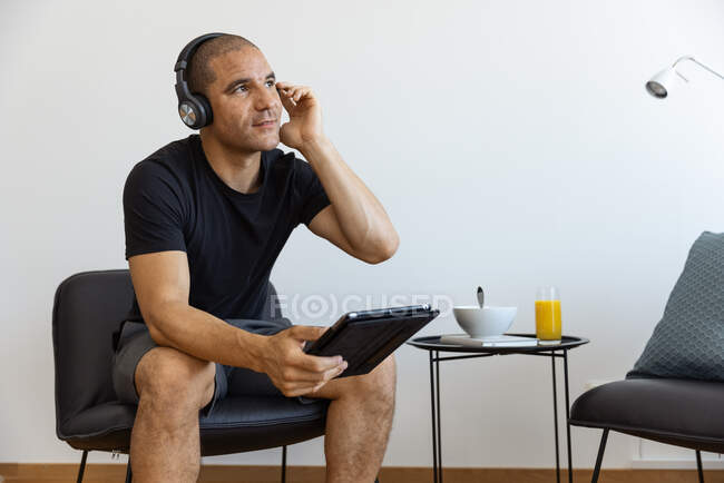 Male in headphones watching video on tablet sitting on chair in the morning at home looking away — Stock Photo