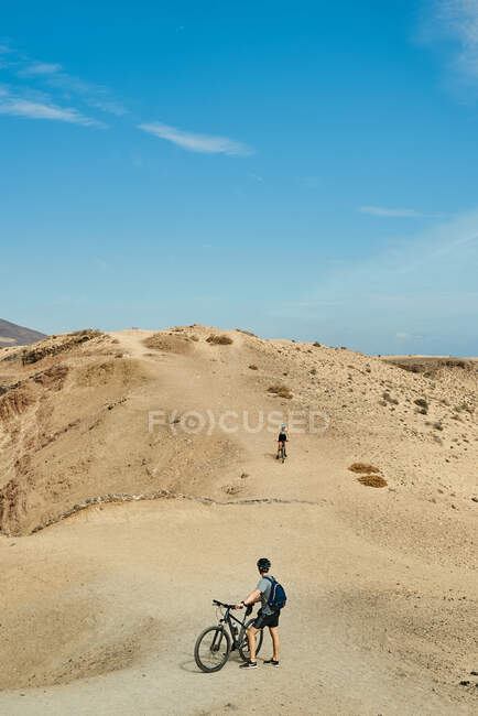 Uomo con zaino e casco in piedi vicino alla bicicletta e guardare l'amico cavalcare su una collina sabbiosa arida nella giornata di sole a Fuerteventura, Spagna — Foto stock