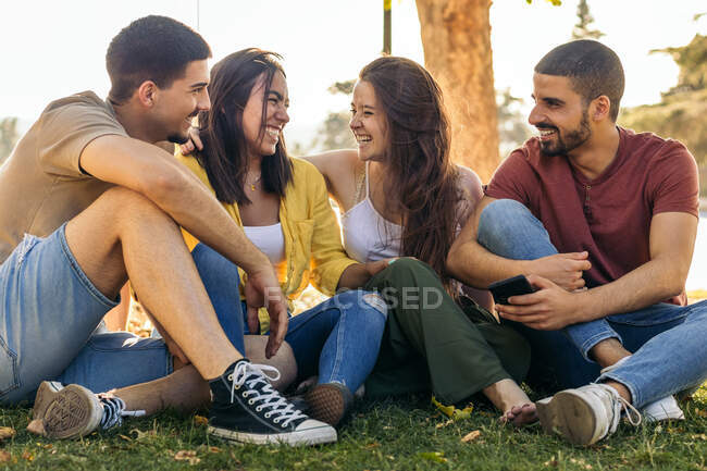 Група молодих чоловіків і жінок посміхаються і розмовляють один з одним, сидячи на траві біля дерева і відпочиваючи в літній день у парку. — стокове фото
