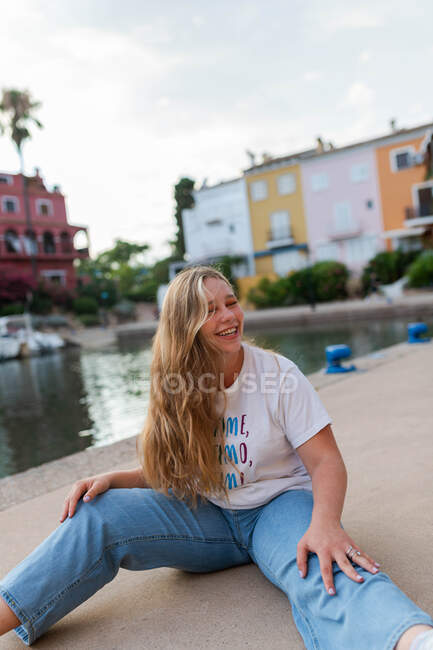 Glückliches stilvolles Weibchen mit langen Haaren sitzt im Sommer auf einem Betondamm in einer exotischen Stadt und blickt in die Kamera — Stockfoto
