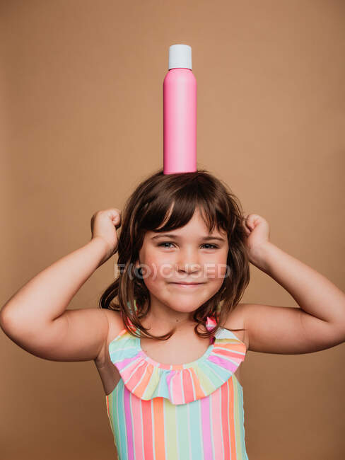 Menina pré-adolescente alegre em maiô de pé com garrafa de creme protetor solar na cabeça em fundo marrom no estúdio e olhando para a câmera — Fotografia de Stock