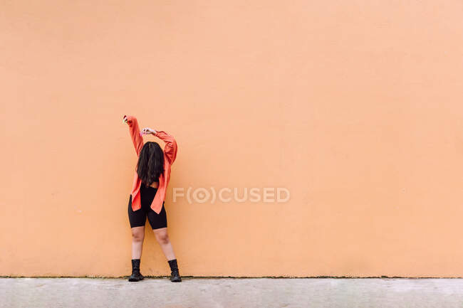 Corps complet de jeune femme anonyme couvrant le visage avec de longs cheveux bruns levant les bras tout en se tenant contre le mur orange — Photo de stock
