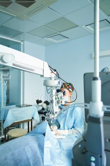 Внимательная женщина-врач в хирургической форме и стерильной маске смотрит через микроскоп во время операции глаза неузнаваемого пациента в больнице — стоковое фото