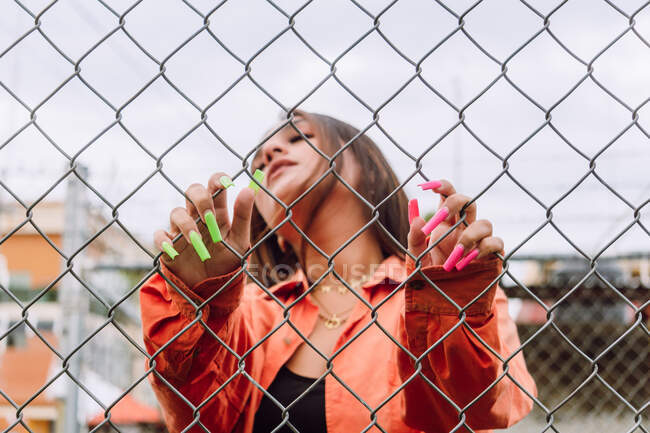 Femme informelle portant une tenue élégante avec de longs ongles brillants tenant une grille métallique avec les yeux fermés sur la rue — Photo de stock