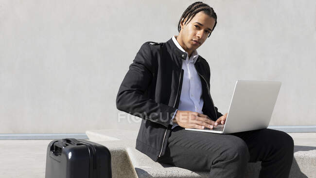 Young ethnic male freelancer with braided hairstyle sitting on bench and typing on laptop keyboard while working on project remotely on urban street — Stock Photo