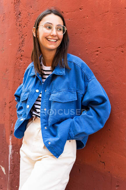 Femme positive dans une tenue élégante regardant loin s'appuyant sur un fond coloré de mur de construction sur une journée ensoleillée dans la rue de la ville — Photo de stock