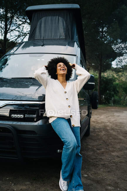 Joyeux jeune voyageuse afro-américaine en vêtements décontractés riant joyeusement tout en s'appuyant sur le camping-car garé dans la nature pendant les vacances d'été — Photo de stock