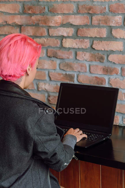 Вид сзади женщина урожая с окрашенными волосами в стильном пальто сидя за столом возле кирпичной стены при использовании ноутбука — стоковое фото