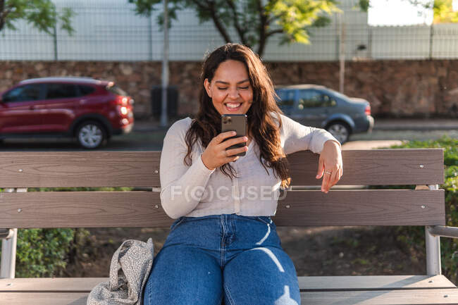 Cheerful young Latin American female student browsing on mobile phone while resting on wooden bench on city street in summer day — Stock Photo
