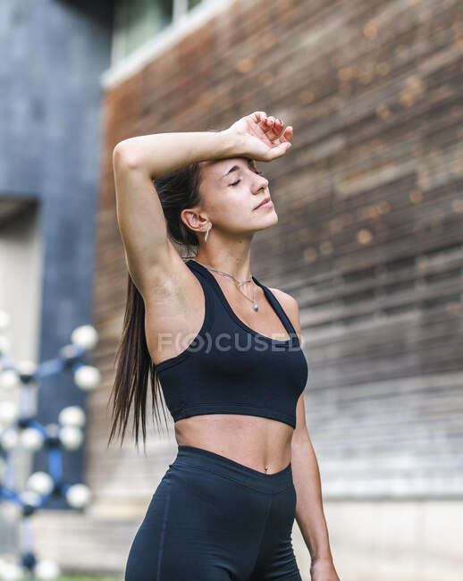 Exhausted female athlete in activewear wiping sweat from forehead with closed eyes during fitness workout on city street — Stock Photo