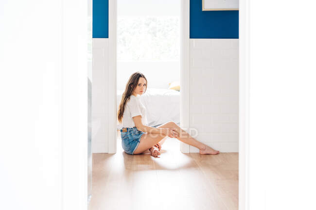 Vista lateral da jovem mulher elegante em shorts jeans e camiseta branca sentada no chão na entrada do quarto em casa e olhando para a câmera — Fotografia de Stock