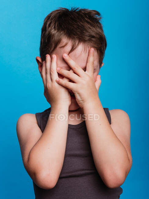 Tímido bonito preteen menino cobrindo rosto com as mãos no vívido azul fundo no estúdio — Fotografia de Stock