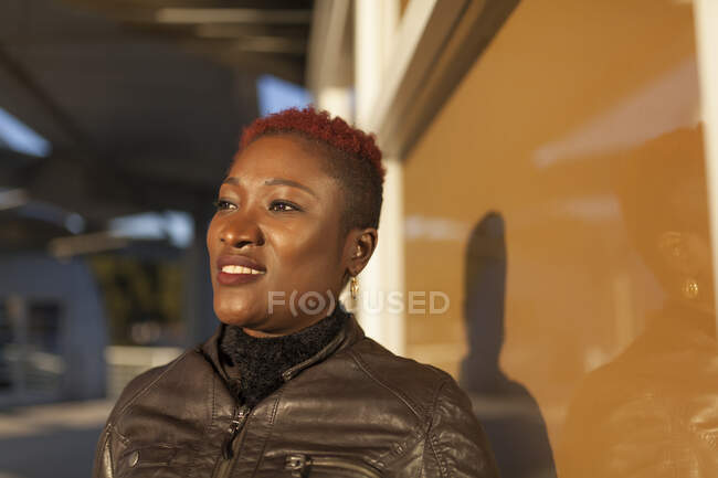Vista lateral da bela mulher afro posando perto da janela e sorrindo enquanto olha para longe em um dia ensolarado — Fotografia de Stock