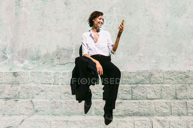 Entzückte Frau in weißem Hemd und Hose sitzt auf Betonrand und hat Videoanruf — Stockfoto