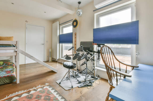 Jeu de tambour électronique placé près de la fenêtre dans une chambre moderne avec lit superposé et table avec chaise dans un appartement de style loft — Photo de stock