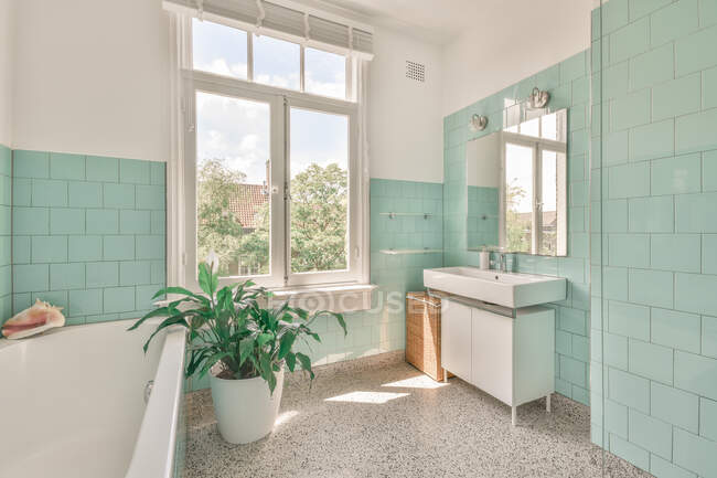 Diseño interior de amplio baño luminoso con ventana y azulejos verdes en las paredes amuebladas con bañera y decoradas con plantas en maceta en casa - foto de stock