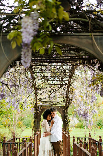 Вид збоку ніжної багаторасової пари, що обіймається, стоячи з очима, закритими під аркою з квітучими фіолетовими квітами вістерії в саду — стокове фото
