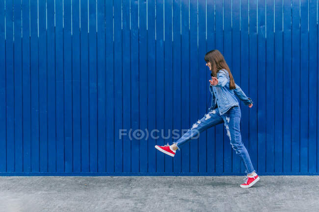 Vista lateral de la vista remota alegre de la mujer en ropa de mezclilla saltando a lo largo de la calle en el fondo del edificio azul - foto de stock
