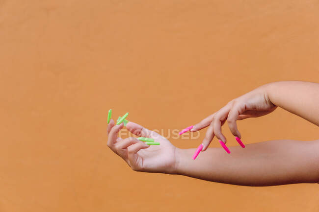 Cosecha femenina anónima con uñas largas cuidadas tocando suavemente la piel de las manos contra el fondo naranja - foto de stock