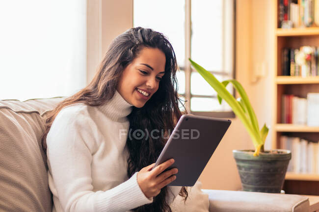 Heureux jeune ethnique femelle navigation sur Internet sur tablette tout en étant assis sur le canapé dans l'appartement — Photo de stock
