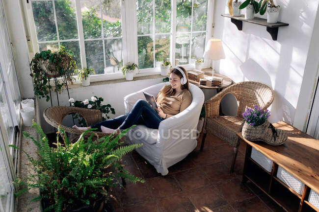 Contenuto femminile in auricolare wireless navigare internet su tablet durante l'ascolto di canzoni in poltrona a casa — Foto stock