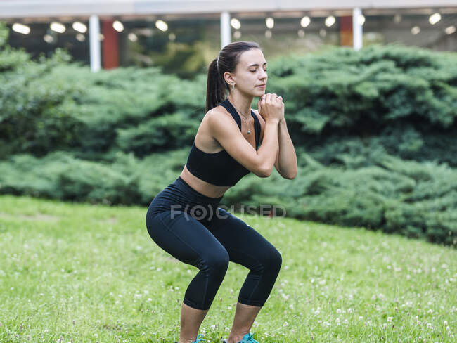 Side view of young sportswoman clasping hands and closed eyes while lunging on grassy lawn during fitness training in summer — Stock Photo